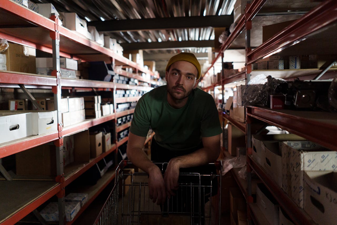 Services Man in green shirt and beanie pushing a cart in a busy warehouse aisle.